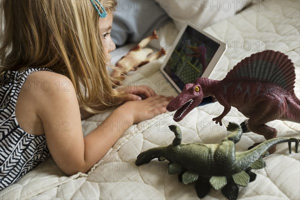 Caucasian girl laying on bed with toy dinosaurs using digital tablet