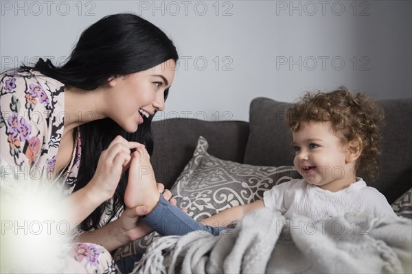 Caucasian mother sitting on sofa playing with toes of son