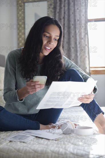 Hispanic woman sitting on bed with cell phone and bills