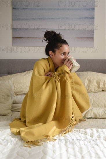 Hispanic woman sitting on bed wrapped in blanket drinking coffee