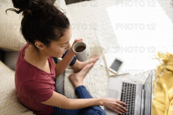 Hispanic woman sitting on bed drinking tea and using laptop