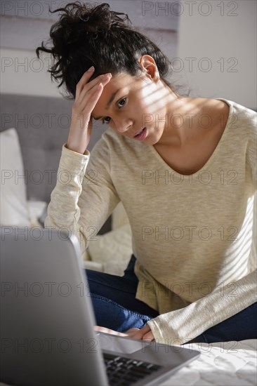 Frustrated Hispanic woman sitting on bed using laptop