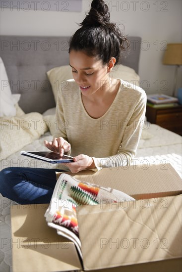 Hispanic woman sitting on bed near box using digital tablet