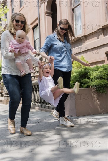 Caucasian mothers swinging daughter on city sidewalk