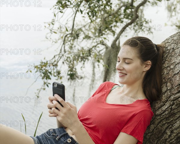 Smiling woman leaning on tree branch and looking at smart phone