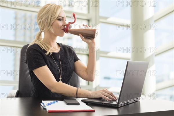 Businesswoman using laptop and drinking protein shake in office