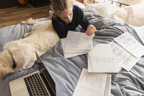 Teenage girl (16-17) lying on bed with dog and doing homework