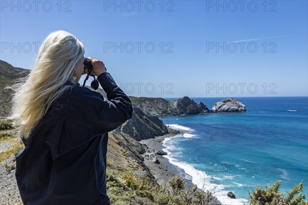 Rear view of senior blonde woman looking at ocean with binoculars on Big Sur coast
