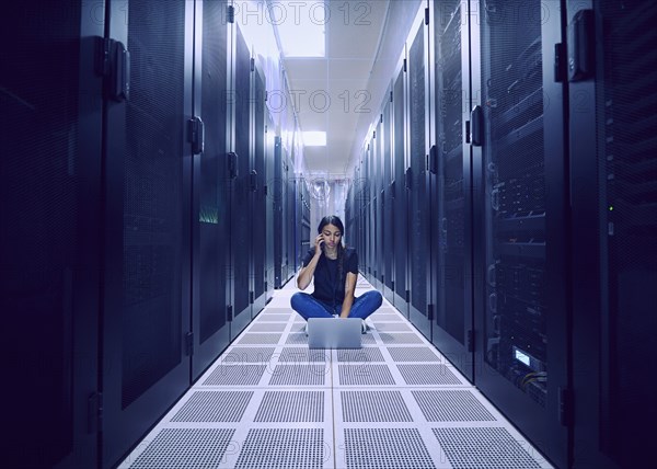 Female technician using laptop and smart phone on floor in server room