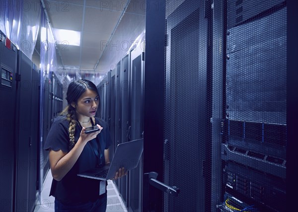 Female technician using smart phone and laptop in server room