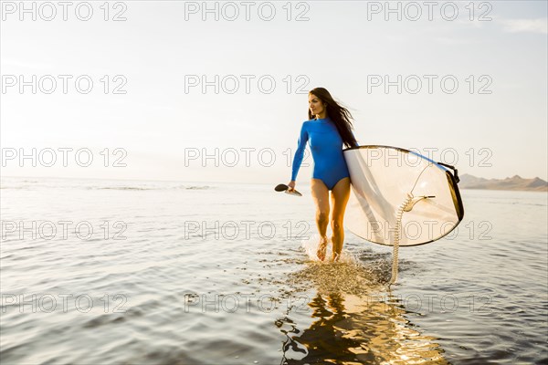 Woman in blue swimsuit carrying paddleboard in lake at sunset