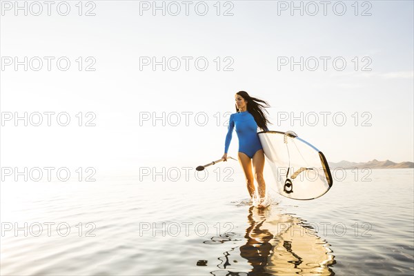 Woman in blue swimsuit carrying paddleboard in lake at sunset