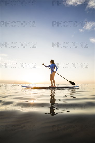 Woman in blue swimsuit paddleboarding on lake at sunset