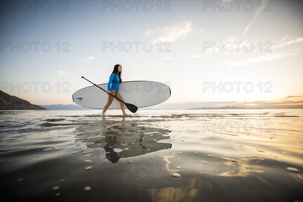 Woman in blue swimsuit carrying paddleboard in lake at sunset