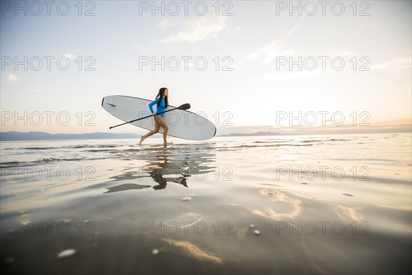 Woman in blue swimsuit carrying paddleboard in lake at sunset