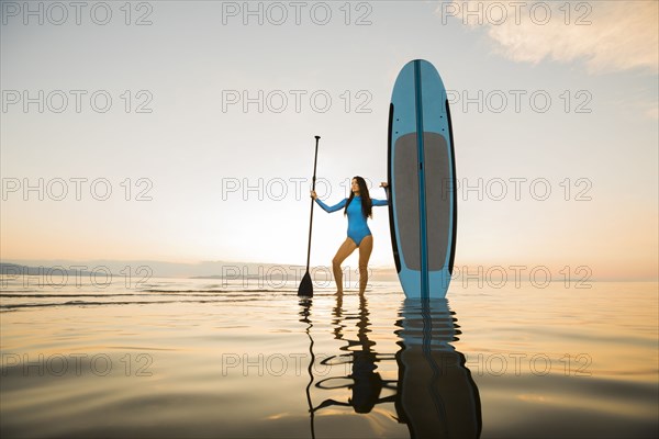 Portrait of woman in blue swimsuit standing with paddleboard in lake at sunset