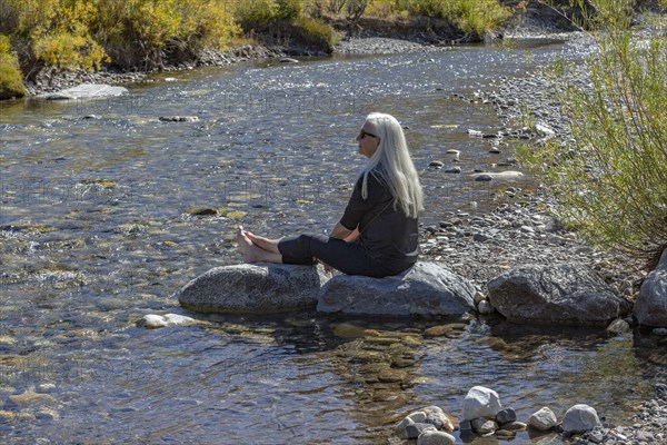 Senior blonde woman sitting on rock by creek