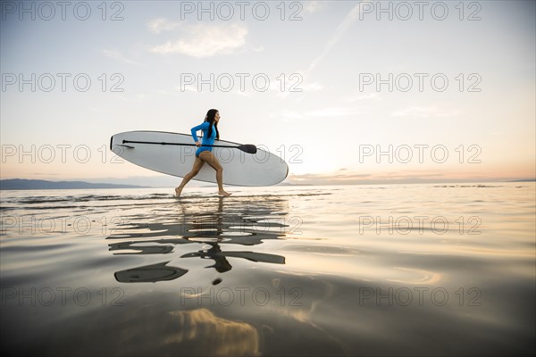 Woman running on beach with paddleboard at sunset