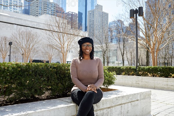 Portrait of smiling woman wearing eyeglasses and beret sitting on wall in city