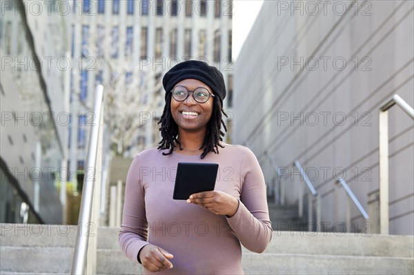 Smiling woman wearing eyeglasses and beret holding digital tablet in city