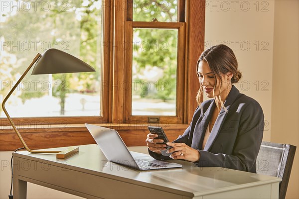 Smiling woman using laptop and smart phone at desk at home