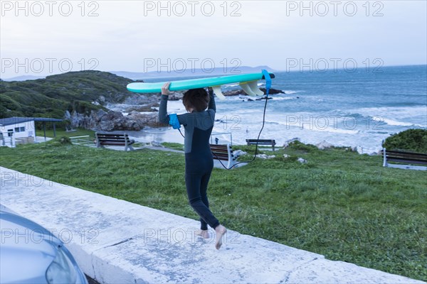 Boy carrying surfboard on head while walking on sidewalk at dusk in Voelklip Beach