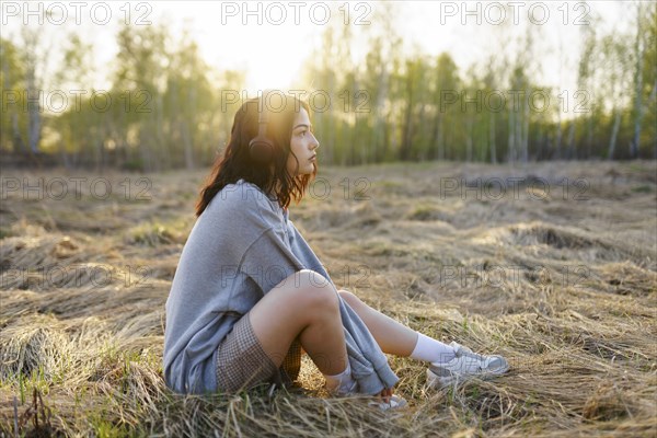 Side view of woman listening to music while sitting on grass in meadow at sunset