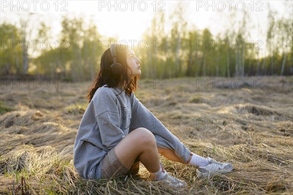 Side view of woman listening to music while sitting on grass in meadow at sunset