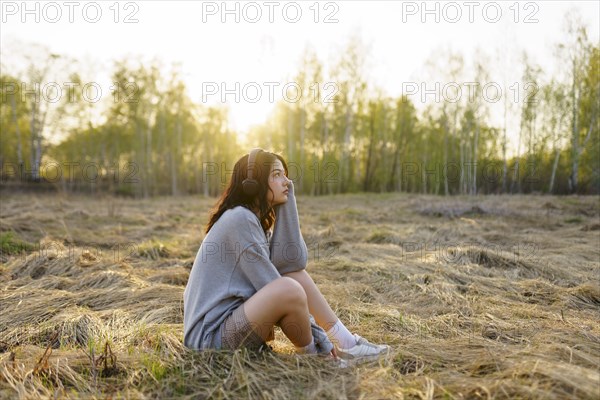 Side view of woman listening to music while sitting on grass in meadow at sunset