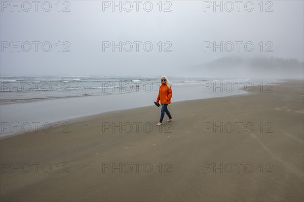 Woman in orange jacket walks along foggy beach