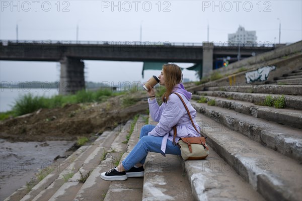 Thoughtful woman sitting on steps and drinking coffee while looking at river
