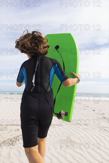 South Africa, Hermanus, Rear view of boy running on beach with body board