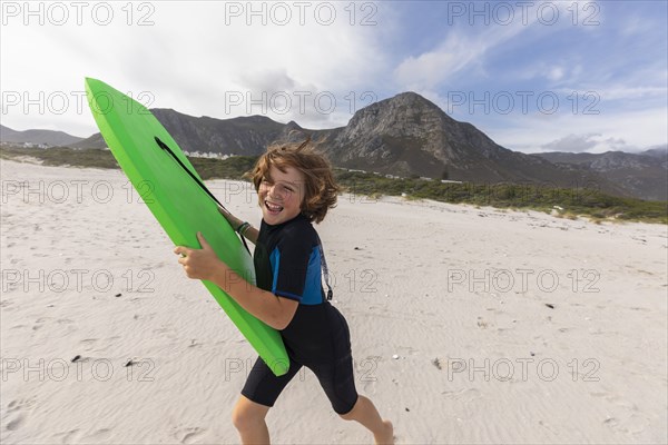 South Africa, Hermanus, Smiling boy running on beach with body board