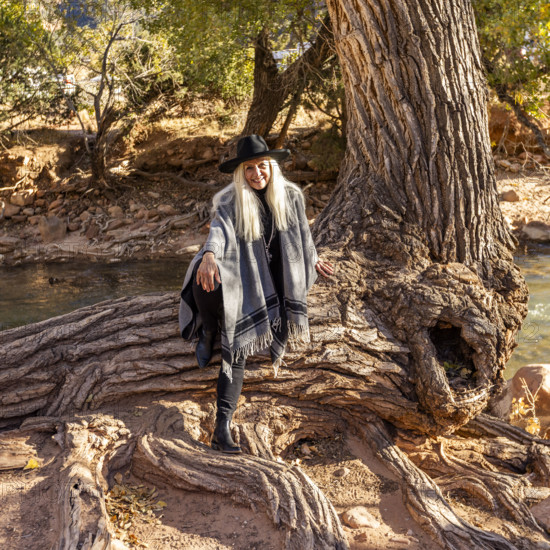 USA, Utah, Zion National Park, Portrait of smiling senior woman in poncho sitting by tree along Virgin River