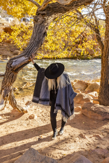 USA, Utah, Zion National Park, Woman in poncho and hat standing by tree along Virgin River