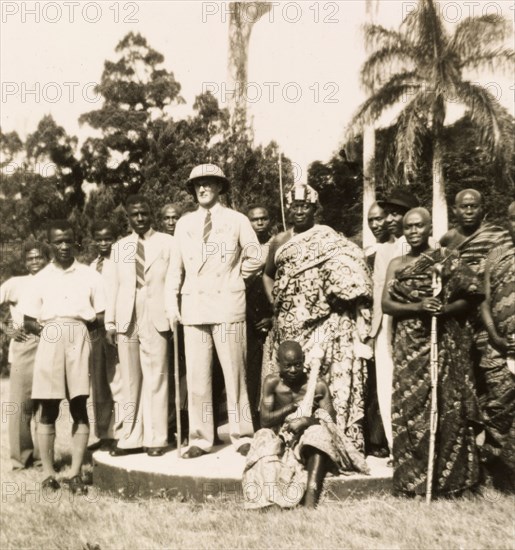 Empire Day in Aburi, 1950. A European colonial officer stands outdoors with an Agona chief, posing for a portrait during celebrations for Empire Day. A group of traditionally dressed Agona men and women, and several African men dressed in Western-style clothing surround the two men. Aburi, Gold Coast (Ghana), 24 May 1950. Aburi, East (Ghana), Ghana, Western Africa, Africa.