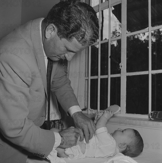 Changing Tubby Block's baby. Tubby Block's baby plays with a hairbrush whilst a man in a jacket and tie, possibly Tubby Block himself, changes it's nappy. Kenya, 30 August 1954. Kenya, Eastern Africa, Africa.