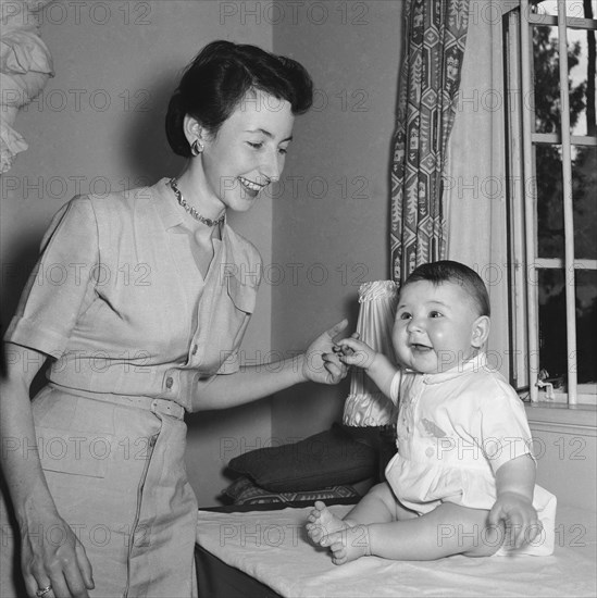Playing with Tubby Block's baby. Interior shot of a smiling woman holding hands with Tubby Block's baby. Kenya, 30 August 1954. Kenya, Eastern Africa, Africa.