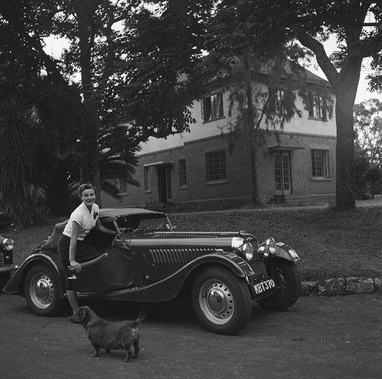 Mrs Douthwaite and car. Mrs Douthwaite and car pictured on the driveway of a large house. There is a small dog in the foreground of the photograph. Kenya, 21 September 1954. Kenya, Eastern Africa, Africa.