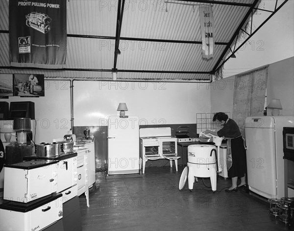 Motor Mart and Exchange Ltd. Interior shot of the Motor Mart and Exchange Ltd display shed at the Royal Show. The photograph features a range of household and kitchen applicances including ovens and fridges. A woman wearing an apron can be seen operating a Thor 230 gyrating washing machine. Nakuru, Kenya, 1 October 1954. Nakuru, Rift Valley, Kenya, Eastern Africa, Africa.