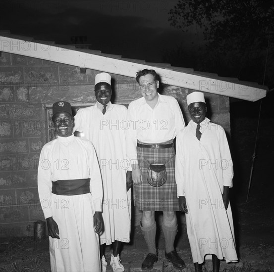 Christening cheer. Exterior night shot of Peter Buchanan and three African servants, all smiling and looking very cheerful. Peter Buchanan is pictured wearing a tartan kilt and sporran. Nairobi, Kenya, 23 October 1954. Nairobi, Nairobi Area, Kenya, Eastern Africa, Africa.