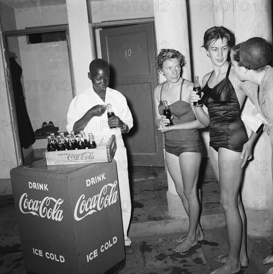 Swim Coca Cola. An African boy opens and distributes bottles of Coca Cola to a group of European girls in swimsuits after their participation in a swimming gala. Kenya, 20 November 1954. Kenya, Eastern Africa, Africa.
