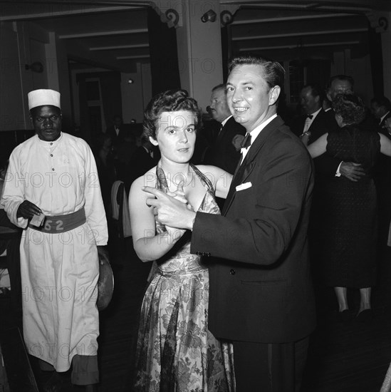 Motor sports dance. A formally dressed couple dancing at the motor sports dance and prize presentation evening. An African waiter wearing a numbered belt appears on the left. Kenya, 26 November 1954. Kenya, Eastern Africa, Africa.