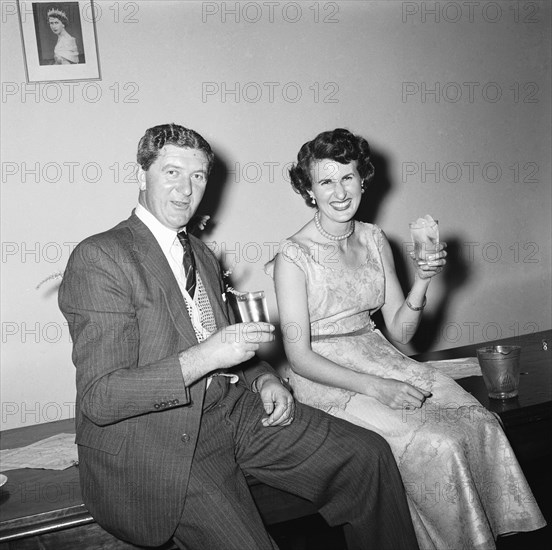 Cheers. Cheerful couple shown seated with drinks below a picture of Queen Elizabeth II at an RAF party buffet. Kenya, 27 November 1954. Kenya, Eastern Africa, Africa.