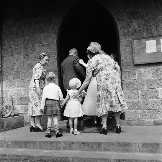 Ushered in. Two smartly dressed women usher a young boy and girl into a church on the occasion of the Macbean wedding. Ngong, Kenya, 18 December 1954. Ngong, Nairobi Area, Kenya, Eastern Africa, Africa.
