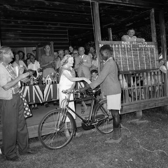 Eldoret prize-giving. An African boy with a bicycle receives congratulations from a European woman at the prize-giving ceremony of the Eldoret race meeting. A crowd of onlookers smile and clap behind a table covered with union jack flags and trophies. Eldoret, Kenya, 27 December 1954. Eldoret, Rift Valley, Kenya, Eastern Africa, Africa.