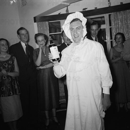 Baby costume. A man wears a fancy dress baby costume at the Archer's New Year party. He holds a bottle of Crawfords old scotch whiskey in one hand and an elephant-shaped baby's rattle in the other. A small crowd of onlookers in the background are clearly entertained. Kenya, 31 December 1954. Kenya, Eastern Africa, Africa.
