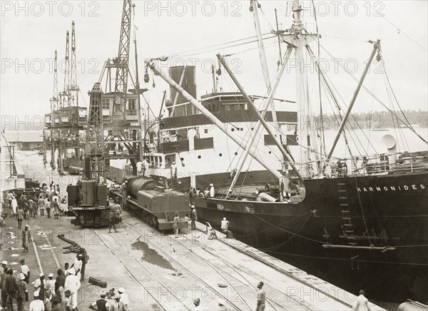 A train unloaded from SS Harmonides. A locomotive boiler and tender have been lowered onto a bogie on the dockside railway at Kilindini harbour by cranes onboard SS Harmonides. A railway steam crane stands by the train. Mombasa, Kenya, circa 1926. Mombasa, Coast, Kenya, Eastern Africa, Africa.