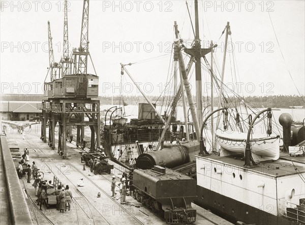 Unloading a train from SS Harmonides. Cranes onboard SS Harmonides hoist a locomotive boiler onto the dockside at Kilindini harbour, where a tender (water tank railcar or wagon) already stands. Mombasa, Kenya, circa 1926. Mombasa, Coast, Kenya, Eastern Africa, Africa.