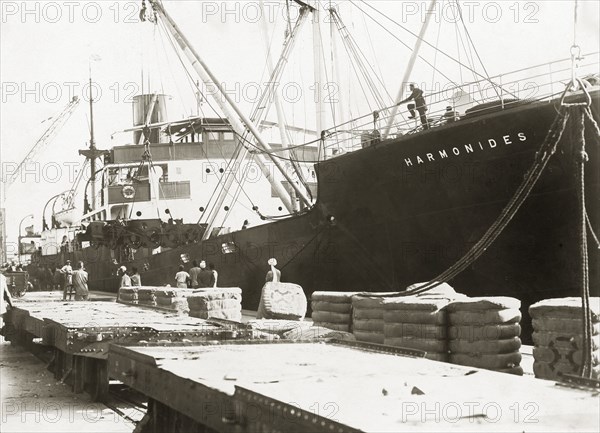 Bogies and SS Harmonides. Cranes onboard SS Harmonides prepare to lower a power bogie onto the dockside railway at Kilindini harbour, where a string of bogies already stand. Mombasa, Kenya, circa 1926. Mombasa, Coast, Kenya, Eastern Africa, Africa.